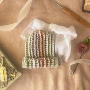 Ribbed baby beanie in sage, clay, and cream tones styled on parchment fabric with crochet hook, vintage book, and lace ribbon — a cozy flat lay highlighting texture and handmade detail.