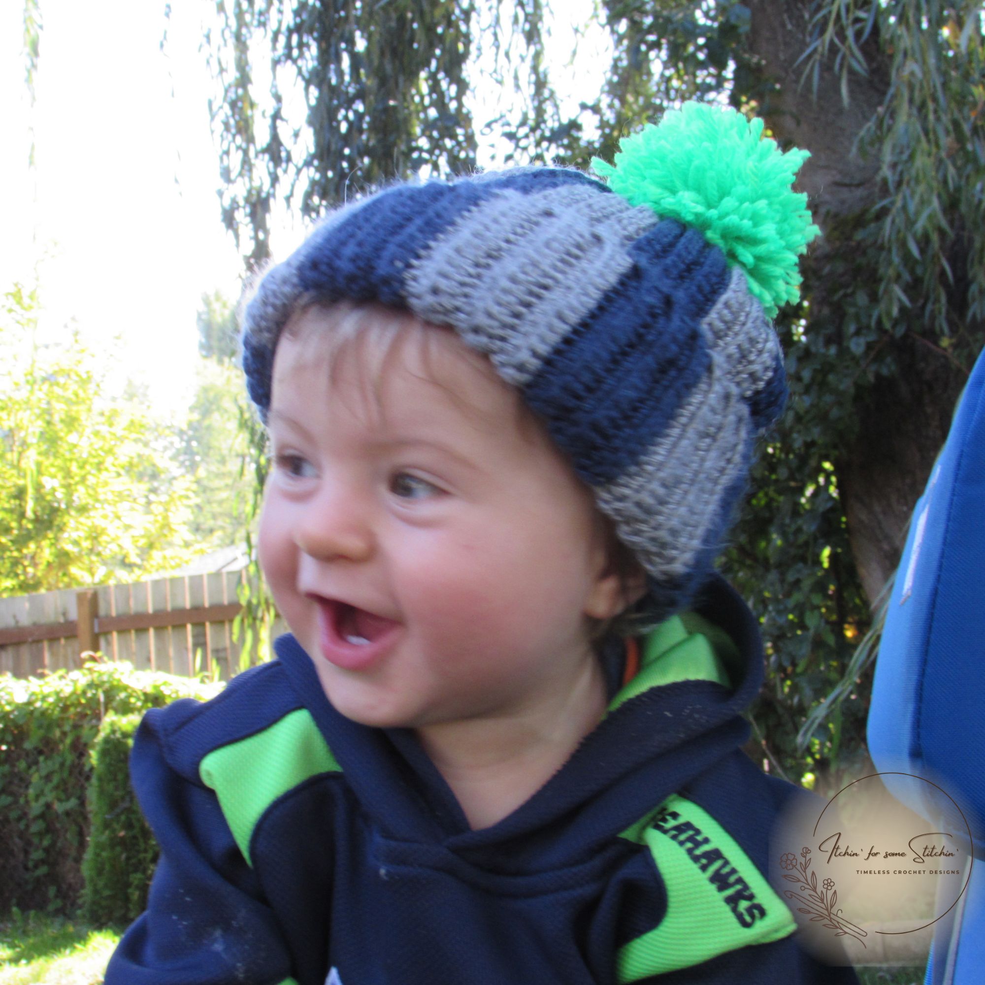 Young child wearing a blue and gray striped ribbed crochet beanie with a green pom-pom, showing the hat’s fit when worn.