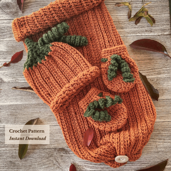 Flat lay of a handmade crochet pumpkin baby set, including an orange ribbed sleep sack, matching hat with green stem and vines, and tiny mittens, styled on a wood surface with autumn leaves. Ideal for fall baby photos and Halloween gifts.