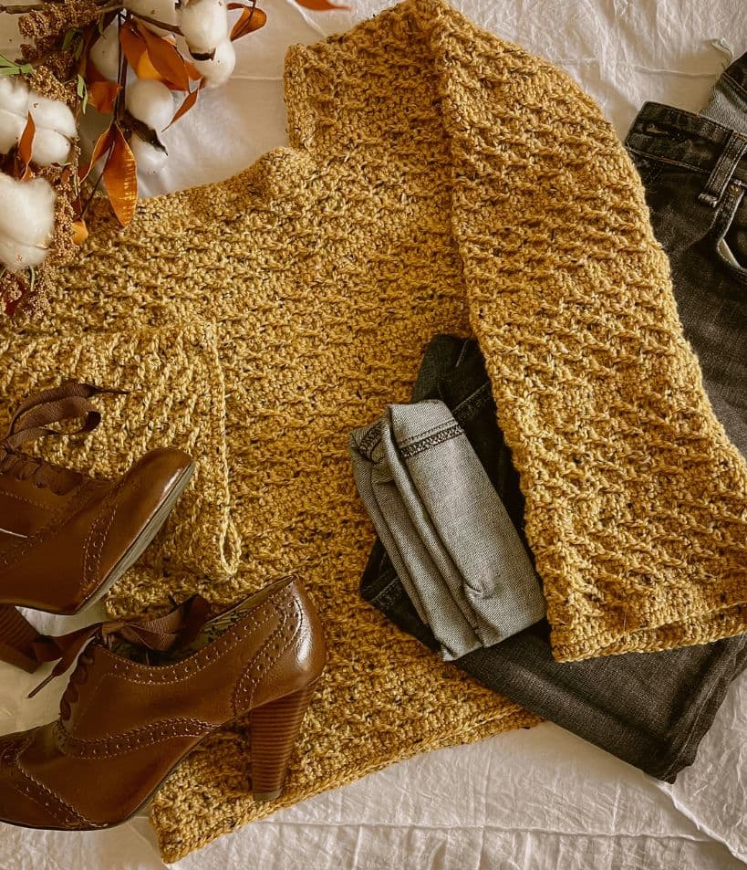 Flat lay of a mustard yellow hand-crocheted sweater paired with vintage brown heels, denim jeans, and a cotton stem bouquet on a white linen background.