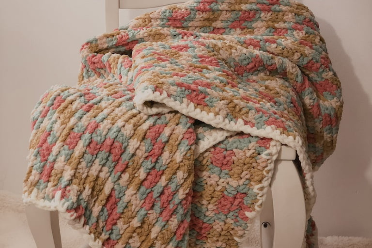 Easy chunky crochet blanket draped over a white vintage chair, featuring plush textured stitches in soft pastel yarn - perfect for beginners. Pattern name is Ly's Blanket.