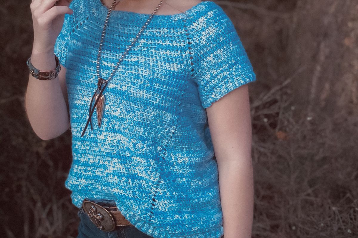 Girl wearing a hand-crocheted blue raglan tee with short sleeves, styled with a wide-brimmed brown hat and western accessories in a rustic outdoor setting.  The top is the Penelope Raglan Tee crochet pattern.