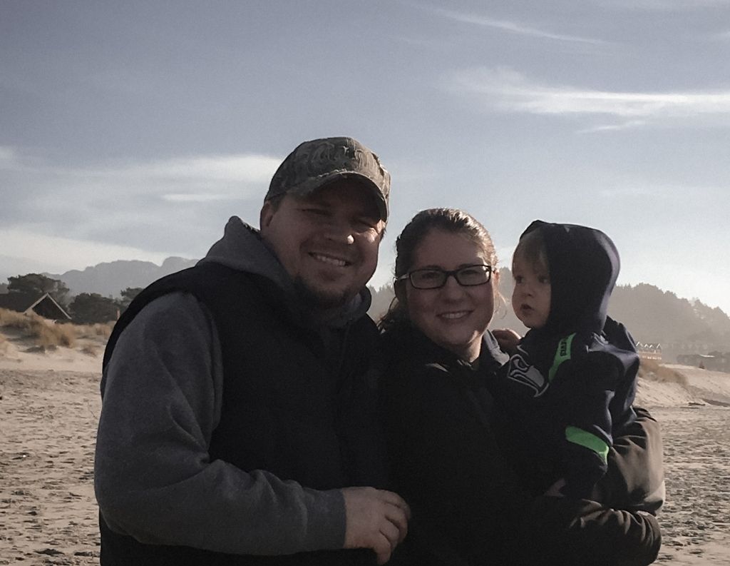 A family of three stands on a beach under a soft, vintage-filtered sky. The mother holds a young child while the father stands beside them, all smiling warmly. The image has a muted, nostalgic tone with a modern vintage French chic aesthetic.