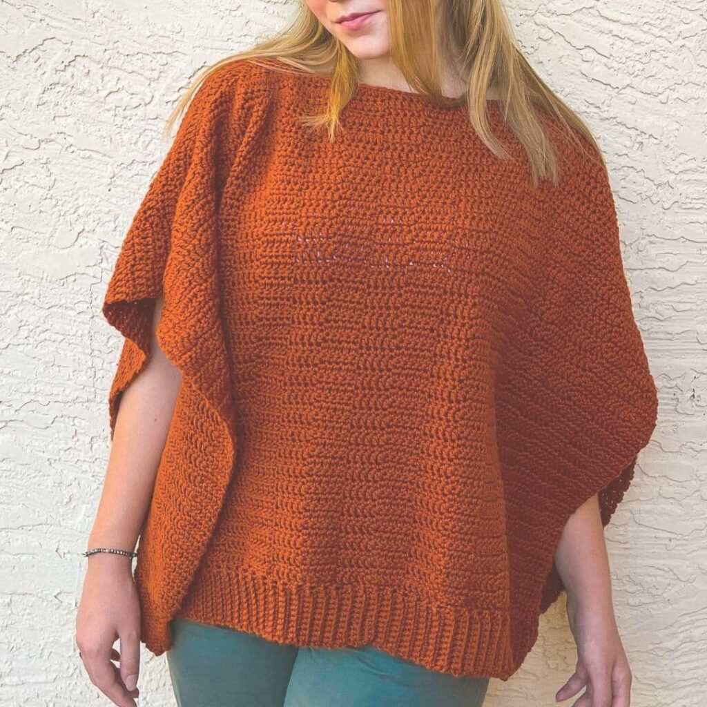 Woman wearing a burnt orange Pennington Poncho, crocheted in textured stitches. Shows completed pattern available through VIP Club.