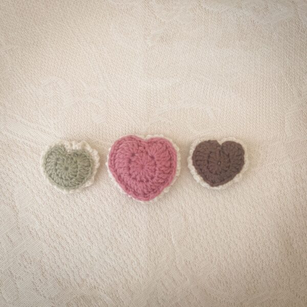 Three crochet puffed heart keepsakes in green, pink, and brown with cream edging, displayed on lace fabric.