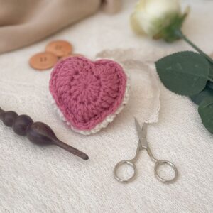 Pink puffed crochet heart keepsake with scalloped edging, yarn needle, scissors, and wooden crochet hook on cream lace background.