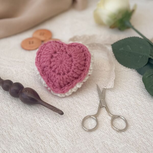 Pink puffed crochet heart keepsake with scalloped edging, yarn needle, scissors, and wooden crochet hook on cream lace background.
