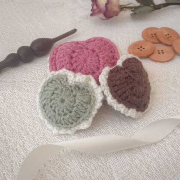 Three handmade crochet puffed hearts in pink, sage green, and brown with cream borders displayed on a lace background. Surrounded by wooden buttons, a crochet hook, dried flower, and satin ribbon, the scene evokes a vintage, handmade charm.