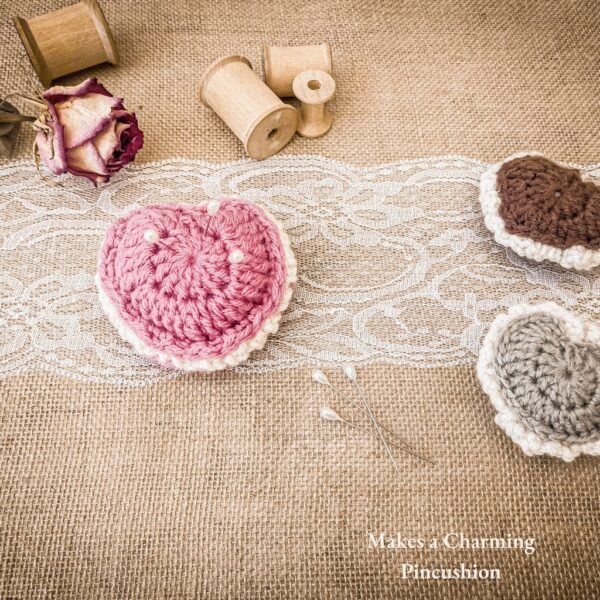Three crochet puffed heart keepsakes in green, brown, and pink, styled with pearl sewing pins vintage bobbins, lace ribbon, and rose on burlap background. Text overlay reads: Makes a Charming Pincushion